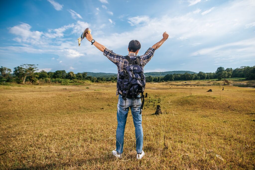 A male traveler with a backpack carrying a map and standing before expanse of vacant land.